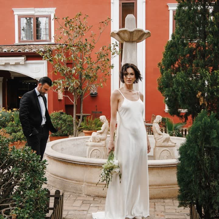 Novia con vestido de seda junto a una fuente en un patio de muros rojos