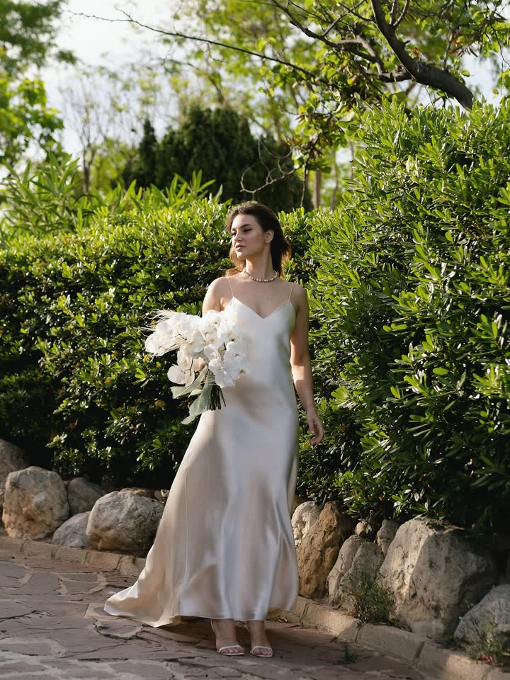 Novia paseando por un camino de jardín con vestido de seda y ramo de orquídeas