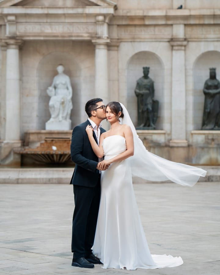 Novios besándose junto a una fuente con estatuas; vestido de seda y velo largo